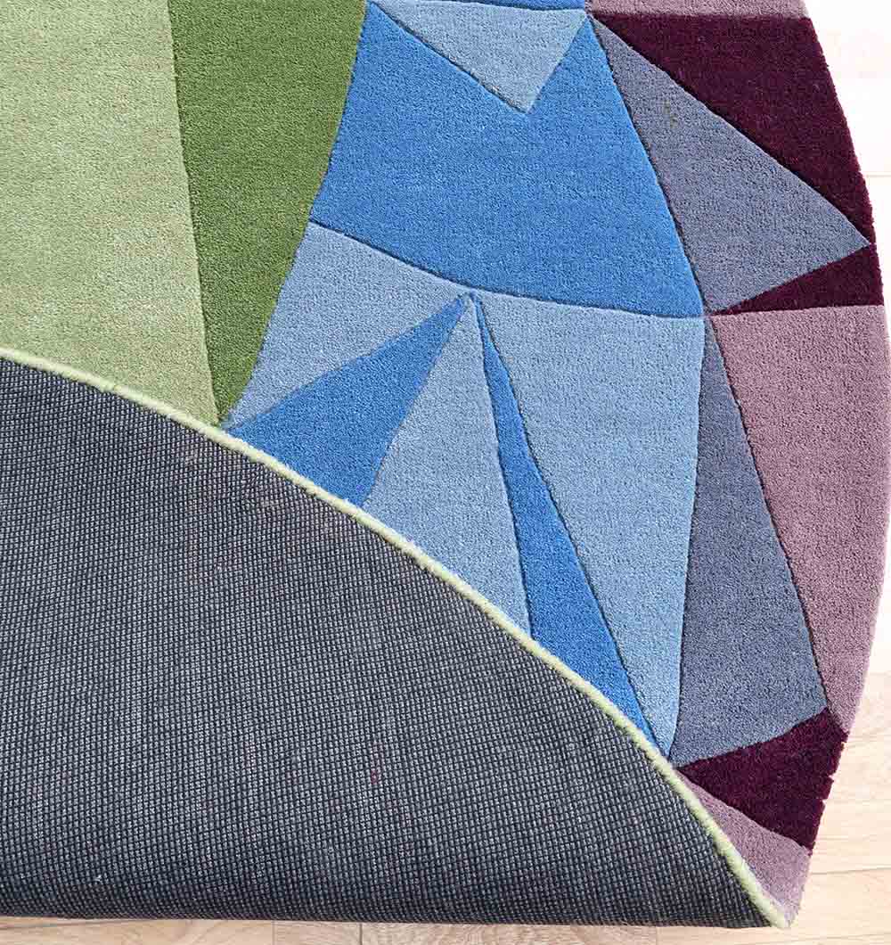 The RAMSHA HOME Multicolor Modern Geometric Round Rug HTW 130 (200cm) features vibrant blue, green, and purple shapes. Partially lifted at the corner, it reveals a textured gray rug pad on a light wood floor for added comfort and style.