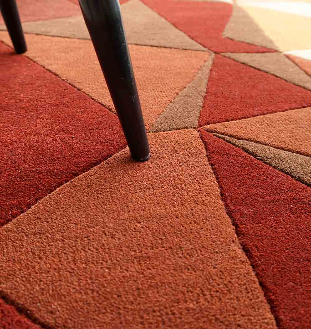 Close-up of RAMSHA HOME’s Multicolor Modern Geometric Round Rug HTW 130 (200cm), featuring a hand-tufted design in red, orange, and brown triangles beneath a black chair leg for a sleek, modern look.