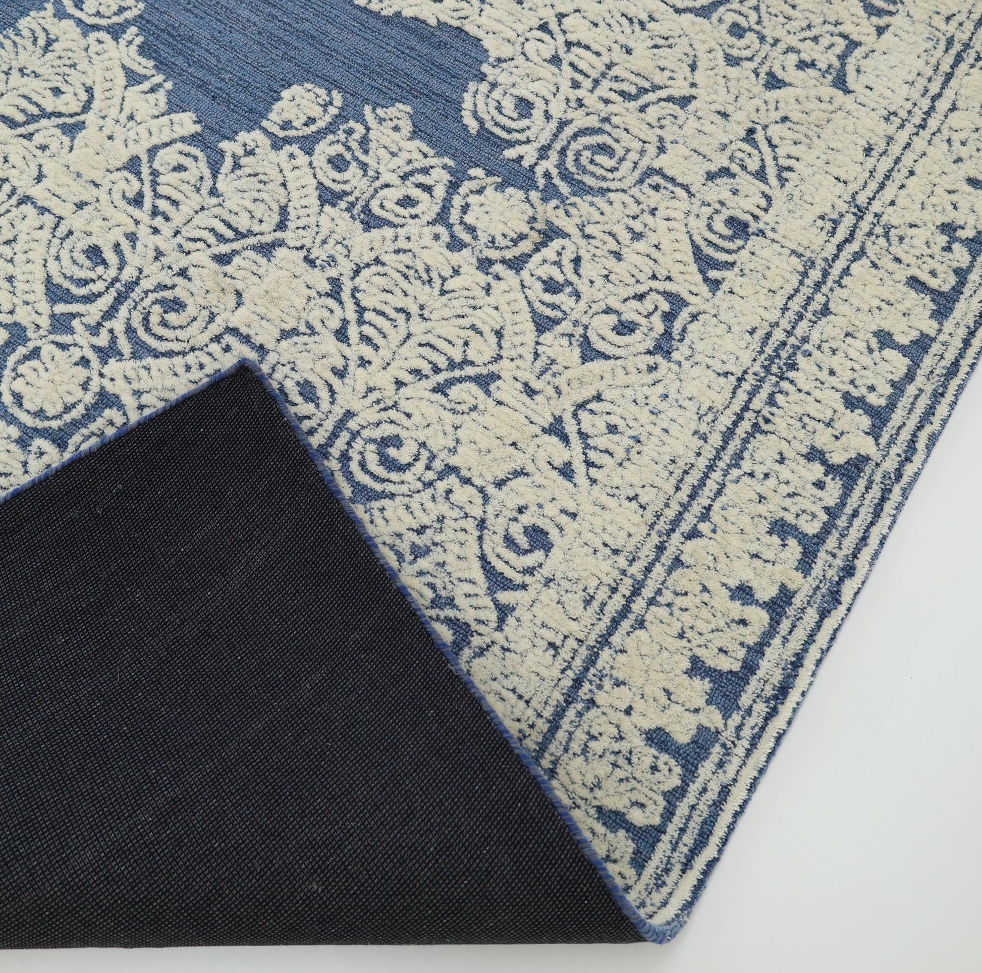 A corner of the Blue Mosaic Handcrafted Luxury Area Rug- HTW 45 by RAMSHA HOME is flipped over, showing the dark underside. This handmade rug displays an ornate floral and scroll border pattern.