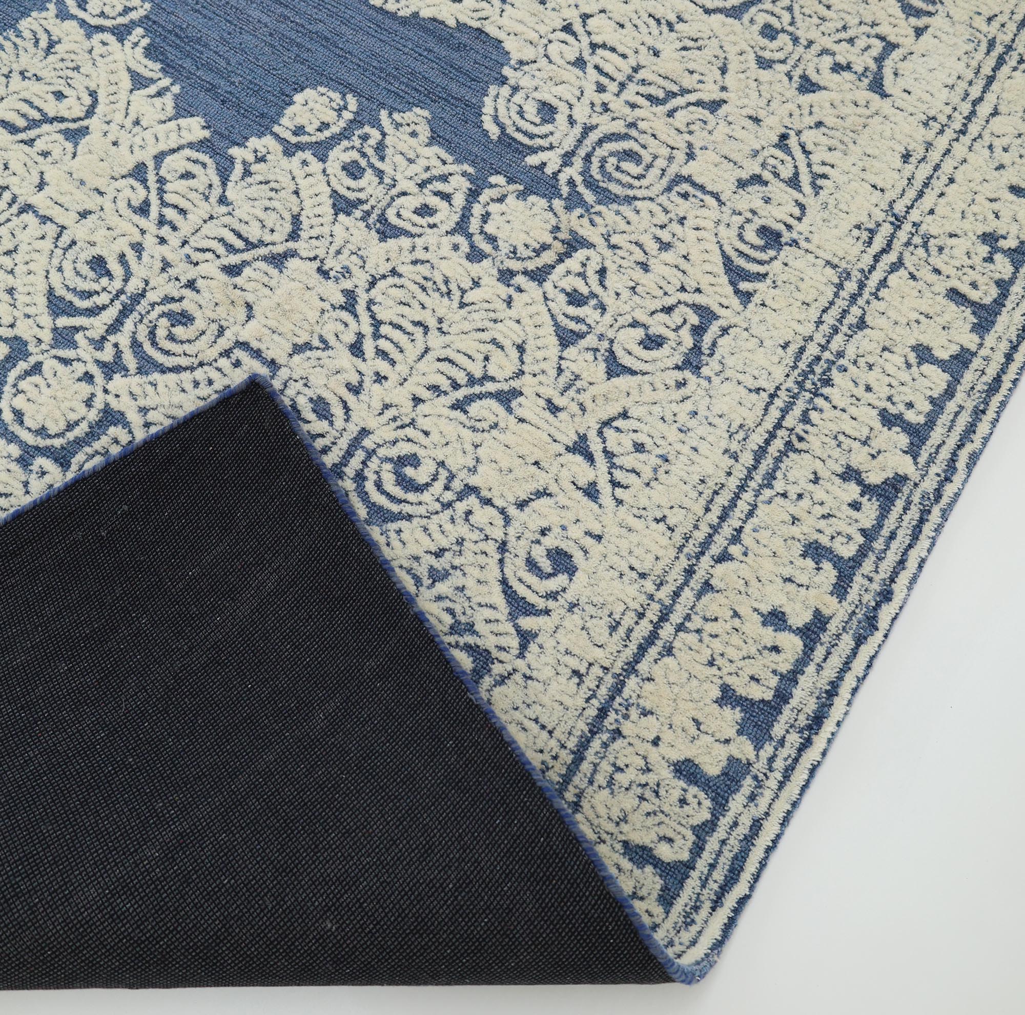 A corner of the Blue Mosaic Handcrafted Luxury Area Rug- HTW 45 by RAMSHA HOME is flipped over, showing the dark underside. This handmade rug displays an ornate floral and scroll border pattern.