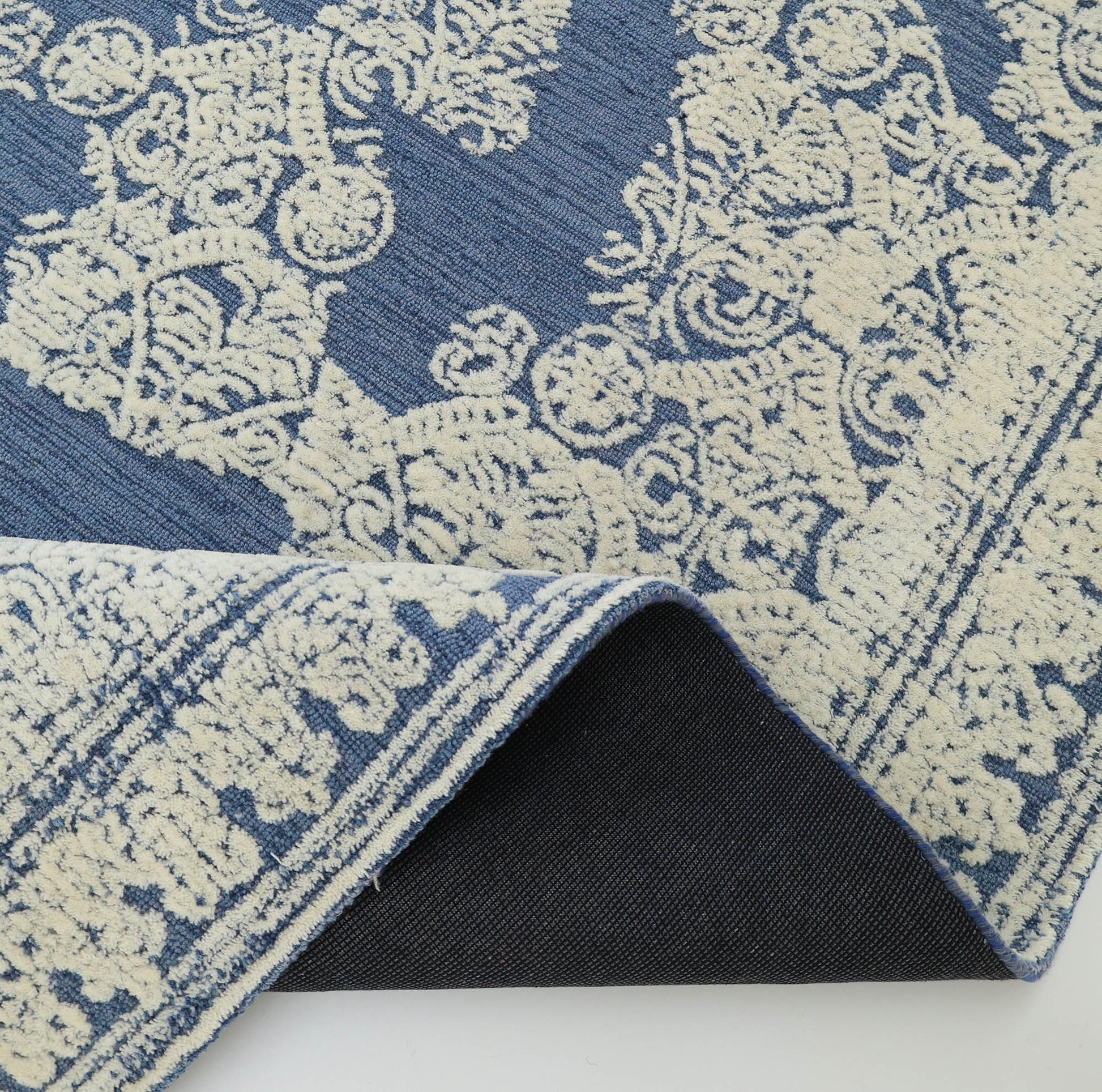 Close-up of the Blue Mosaic Handcrafted Luxury Area Rug- HTW 45 by RAMSHA HOME, featuring a blue distressed design with cream floral motifs and a folded corner revealing its black textured non-slip backing.