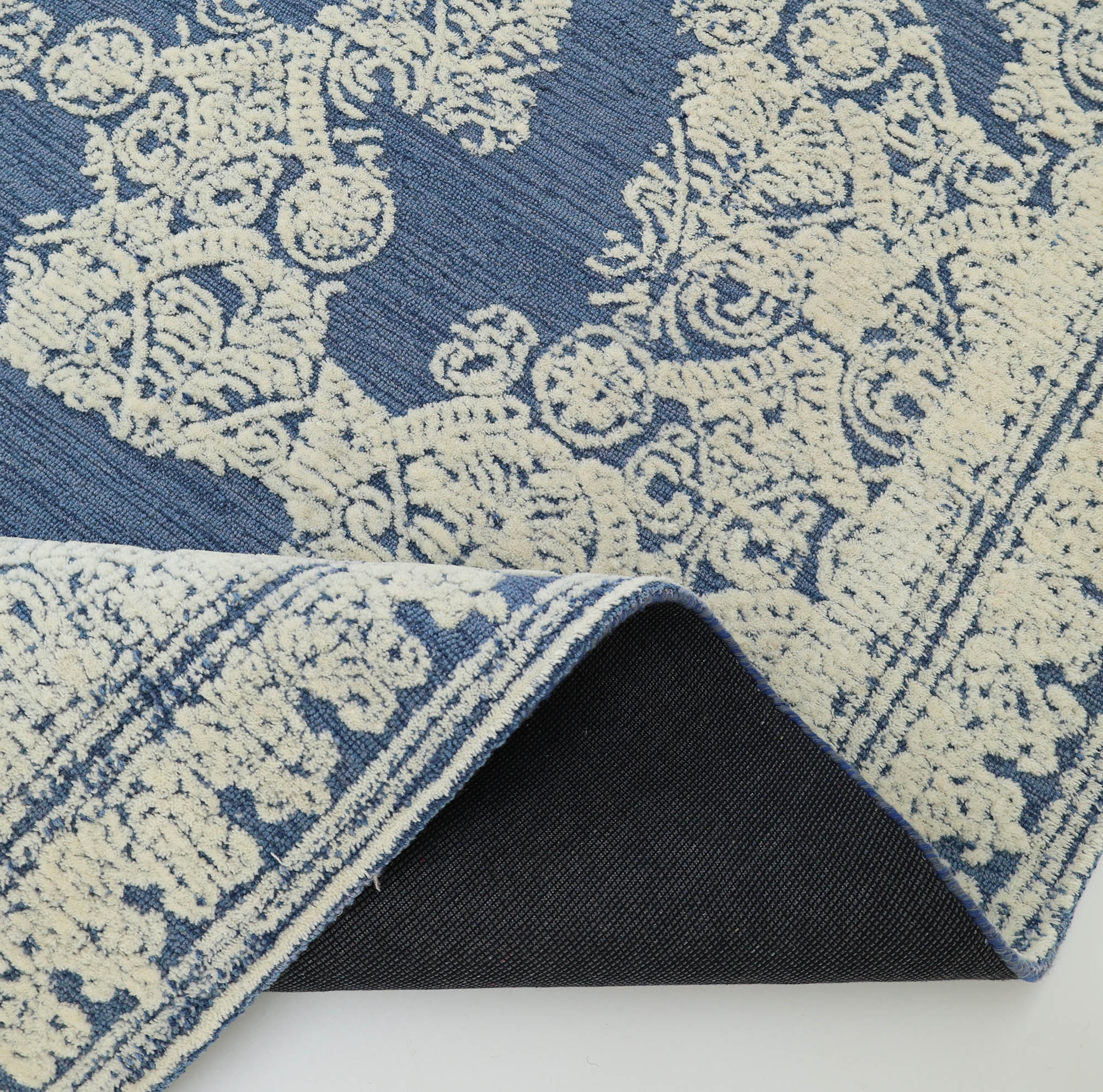 Close-up of the Blue Mosaic Handcrafted Luxury Area Rug- HTW 45 by RAMSHA HOME, featuring a blue distressed design with cream floral motifs and a folded corner revealing its black textured non-slip backing.