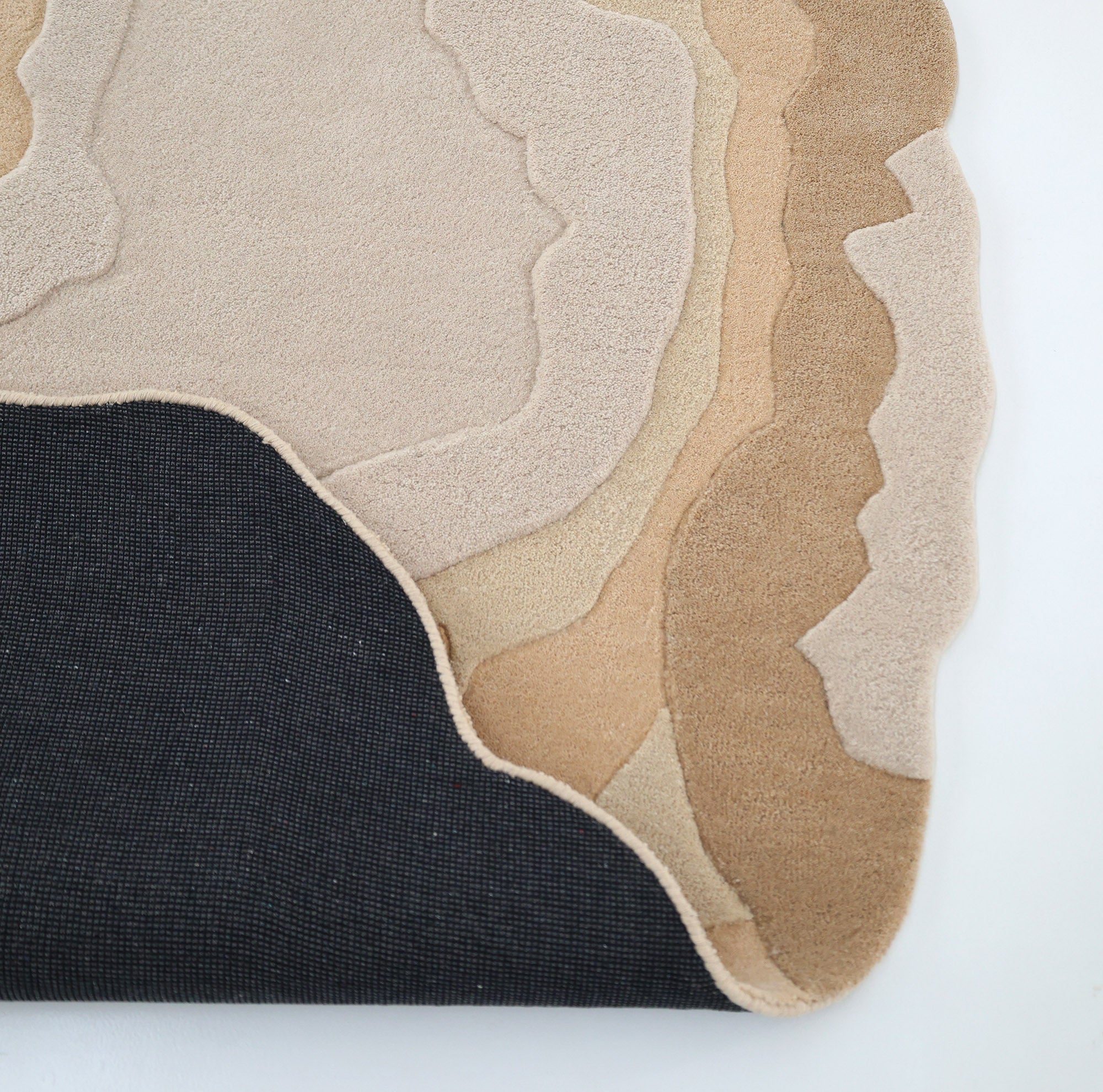 The RAMSHA HOME Contemporary Flow Area Handmade Rug HTWV-153 features layered, wavy shapes in cream, beige, and tan with an abstract border, shown partially folded over a black textured backing against a white background.