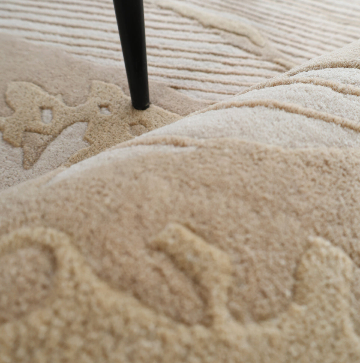 Close-up of the Abstract Beige Rug – Neutral Textured Modern Area Rug with Organic Pattern (HTW 156) by RAMSHA HOME, showcasing its raised, organic patterns in beige tones. A black furniture leg stands on the rug, highlighting the intricate texture and detailed craftsmanship of the carpet surface.