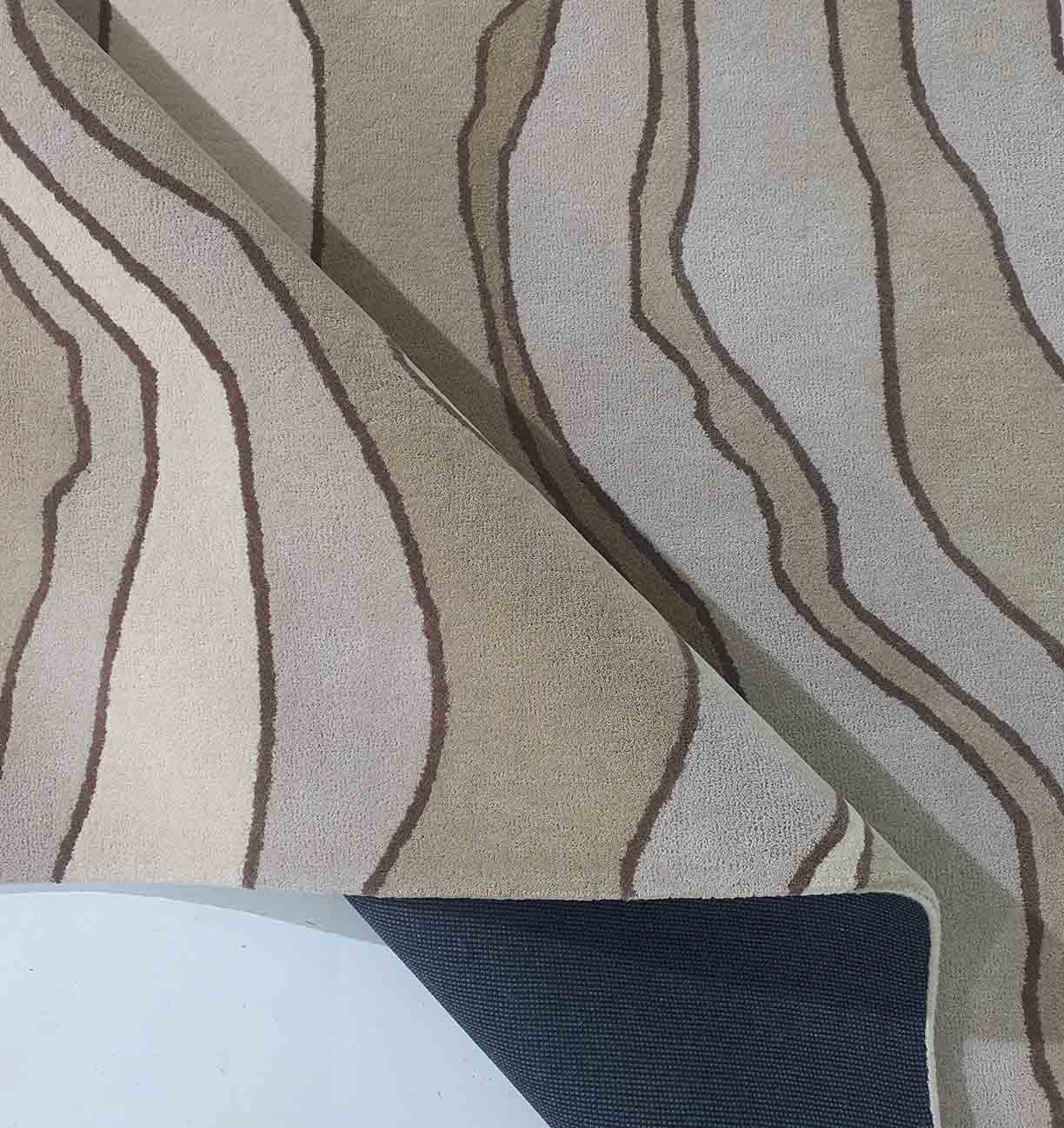 A close-up of the RAMSHA HOME Beautiful Home Decor Handmade Carpet – HTW-66, a hand-tufted beige rectangle rug with wavy brown lines, partially flipped to show its gray underside and backing. The pattern evokes natural wood grain or flowing lines.
