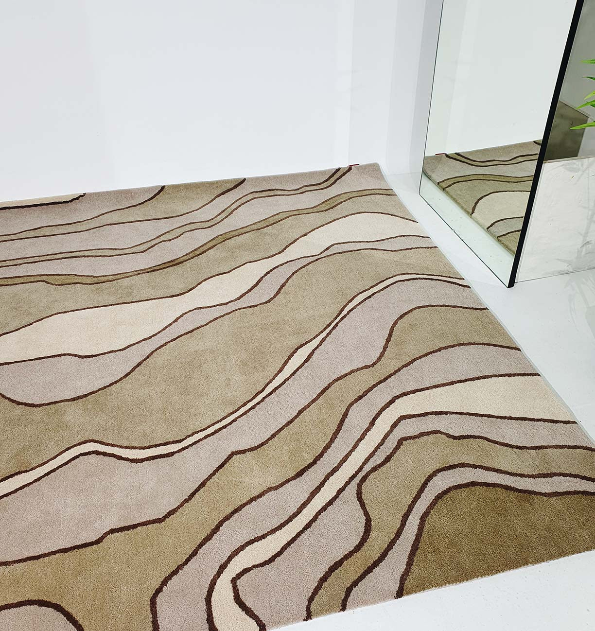 The RAMSHA HOME Beautiful Home Decor Handmade Carpet – HTW-66, featuring beige and brown wavy abstract lines, sits on a white floor beside a large mirror that reflects the stylish hand-tufted rug and part of a green plant.