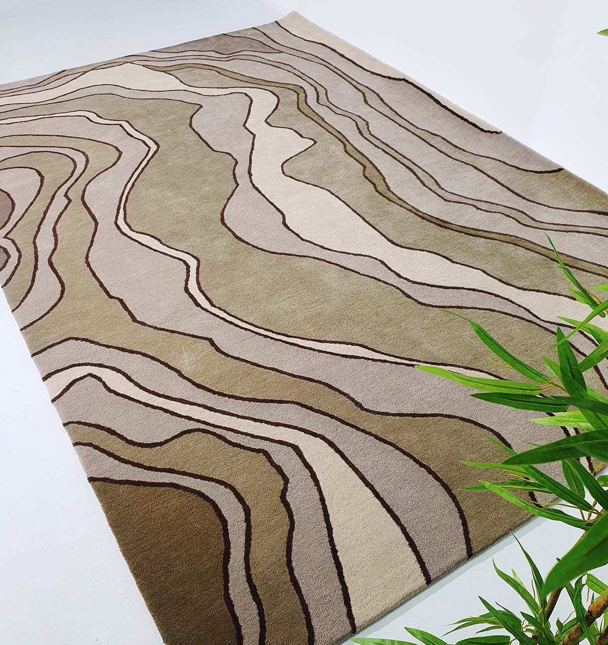 The RAMSHA HOME Beautiful Home Decor Handmade Carpet – HTW-66 features wavy abstract lines in brown, beige, and tan. This hand tufted rug is shown on a light floor with a green leafy plant visible at the bottom right corner.