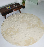 The RAMSHA HOME Round Home Decor Handmade Carpet – HTW-67, hand-tufted from wool with textured crater-like patterns, lies on a light floor beside a wooden coffee table with magazines and a black potted plant.