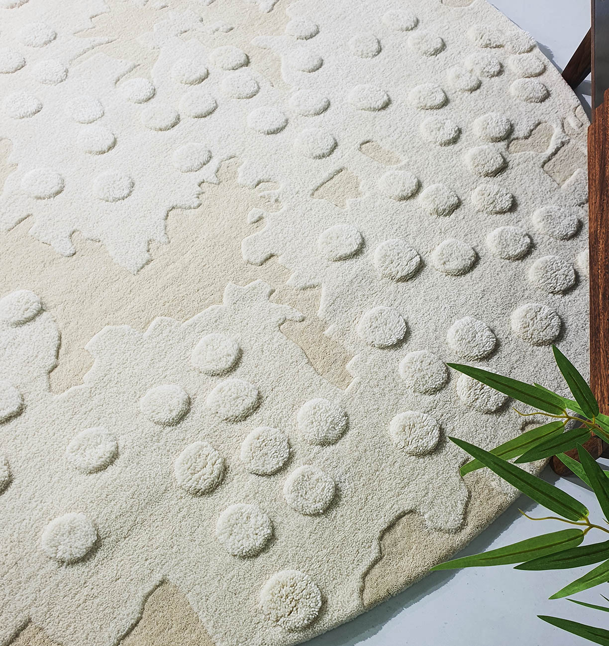 The RAMSHA HOME Round Home Decor Handmade Carpet – HTW-67 in beige and cream with raised circles sits on a light floor, accented by a green plant and a wooden furniture leg in the corner.