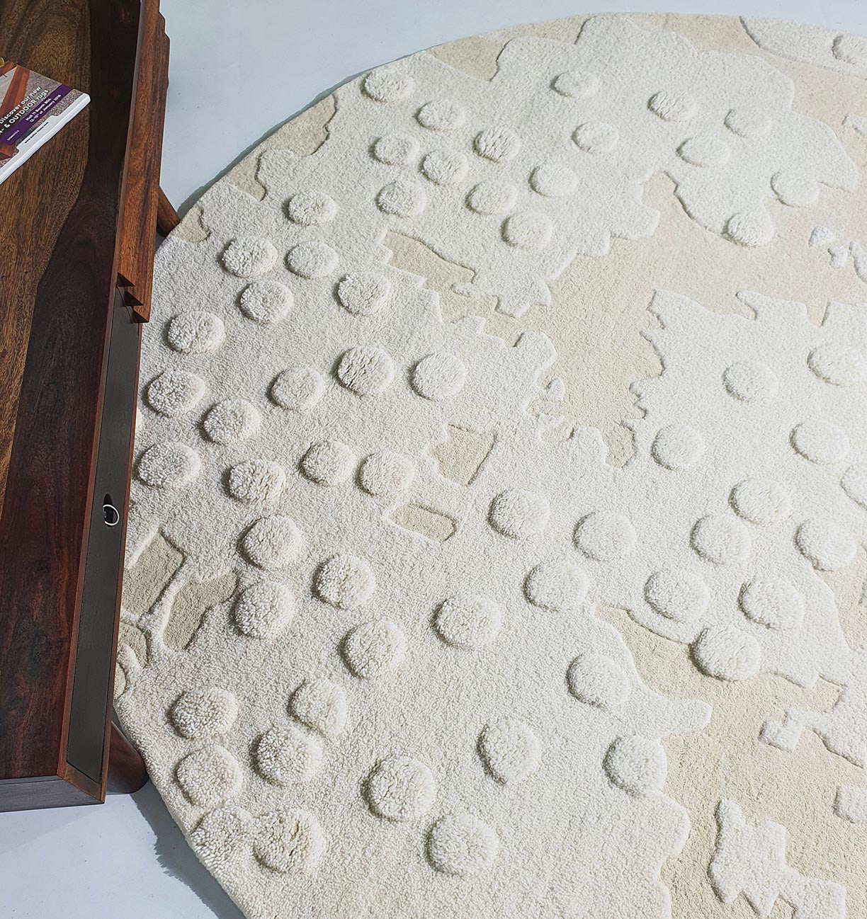 The RAMSHA HOME Round Home Decor Handmade Carpet – HTW-67, a cream-colored hand-tufted wool rug with raised circular and irregular shapes, lies on a light floor beside a dark wooden table holding a magazine.