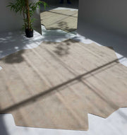 The Stunning Off Flower Shape Handmade Rug - HTW 77 (200 cm) by RAMSHA HOME, in beige wool, lies on a white floor near a potted bamboo and tall mirror, which reflects the modern hand-tufted rug as sunlight creates gentle shadows across the scene.