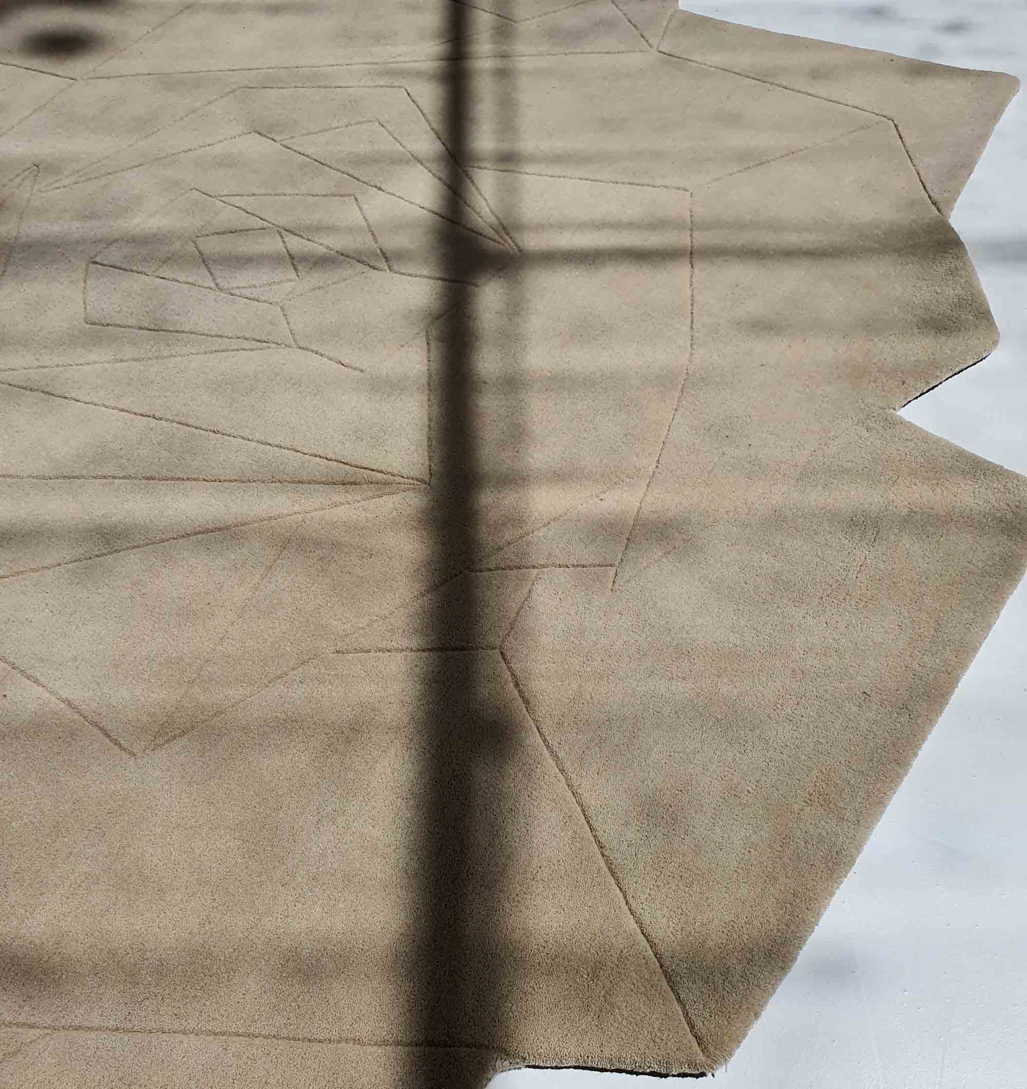 The Stunning Off Flower Shape Handmade Rug - HTW 77 (200 cm) by RAMSHA HOME, in beige with geometric and subtle line patterns, rests on a light floor as diagonal window shadows cross over it.