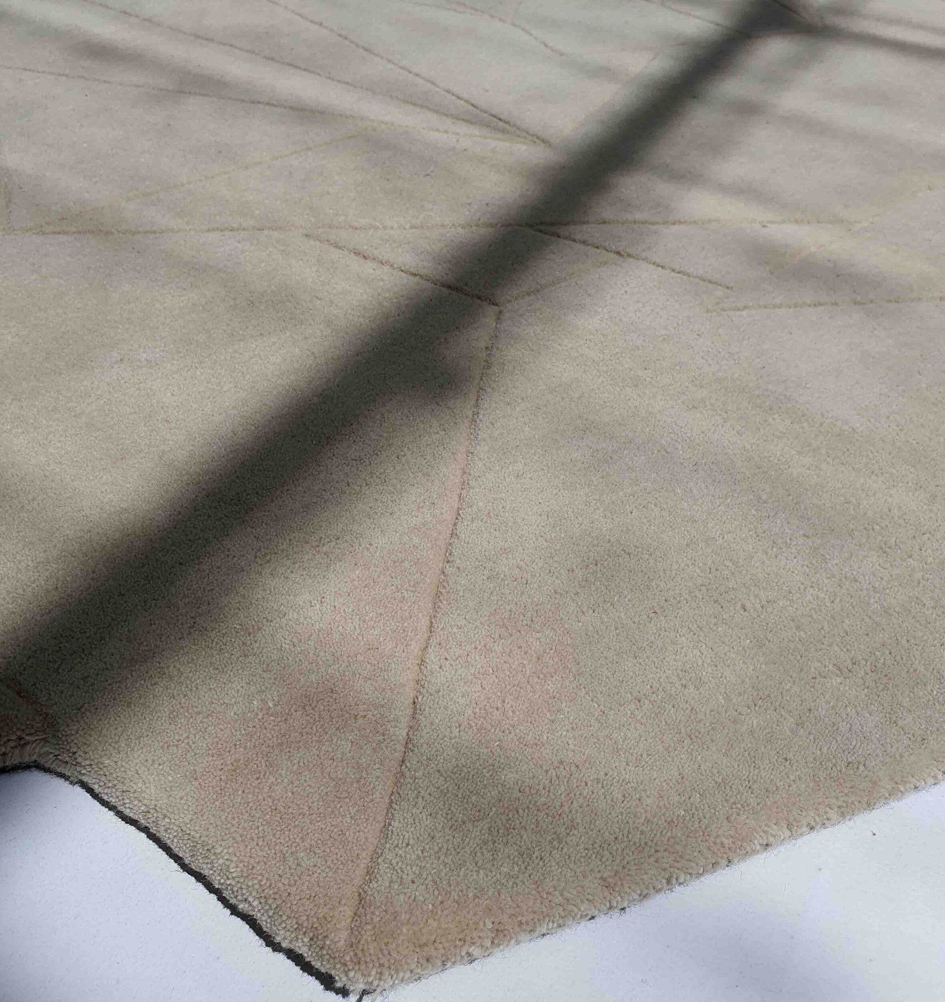A close-up of the RAMSHA HOME Stunning Off Flower Shape Handmade Rug - HTW 77 (200 cm) shows light beige tones with subtle geometric lines and intersecting shadow patterns, with the rug’s edge visible in the corner.