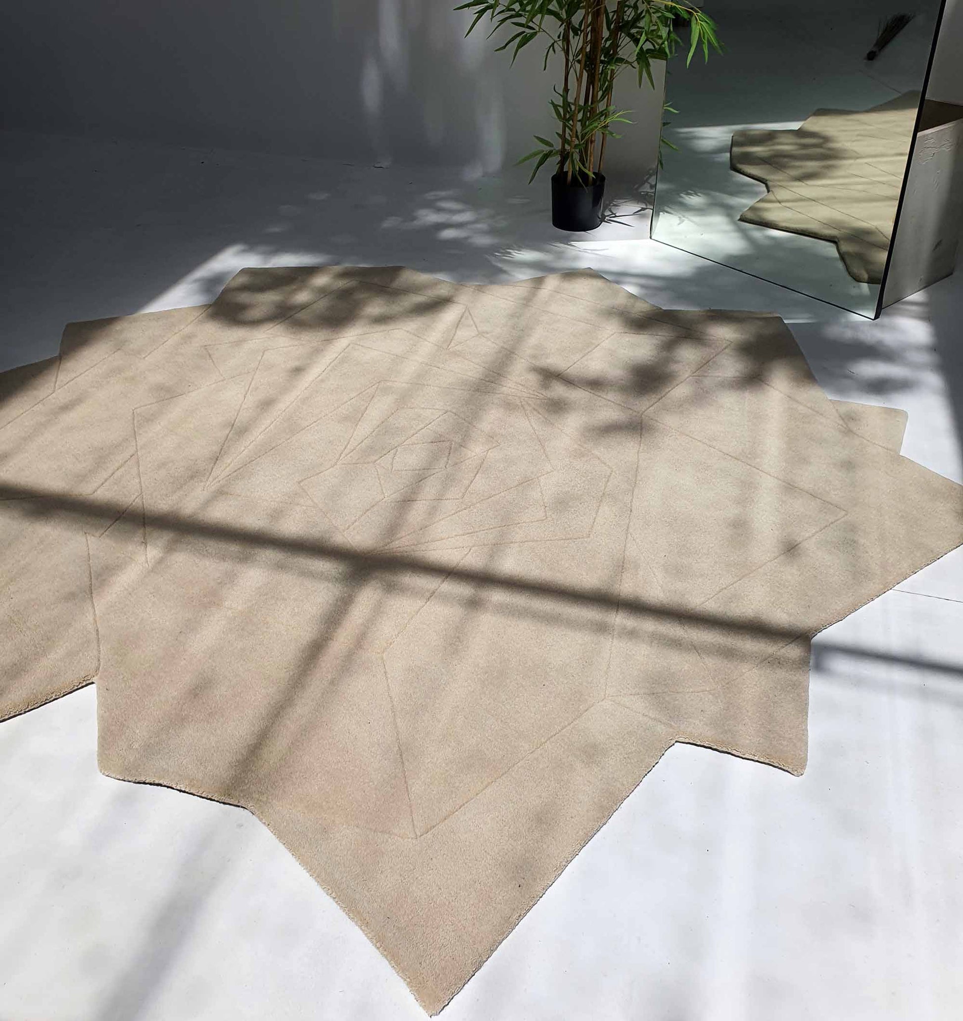The RAMSHA HOME Stunning Off Flower Shape Handmade Rug - HTW 77 (200 cm) features a modern, irregular beige design. Placed on a white floor near a mirror and potted plant, leaf and window shadows fall beautifully across the unique area rug.