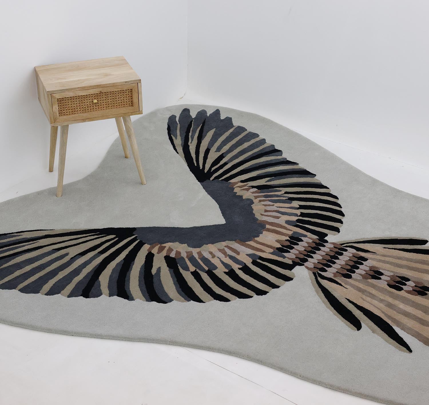 A light wooden nightstand with a rattan drawer stands beside the RAMSHA HOME Unshape Handmade Multi Color Rug HTW-82, which features a large stylized bird in black, brown, and gray on white walls and floor.