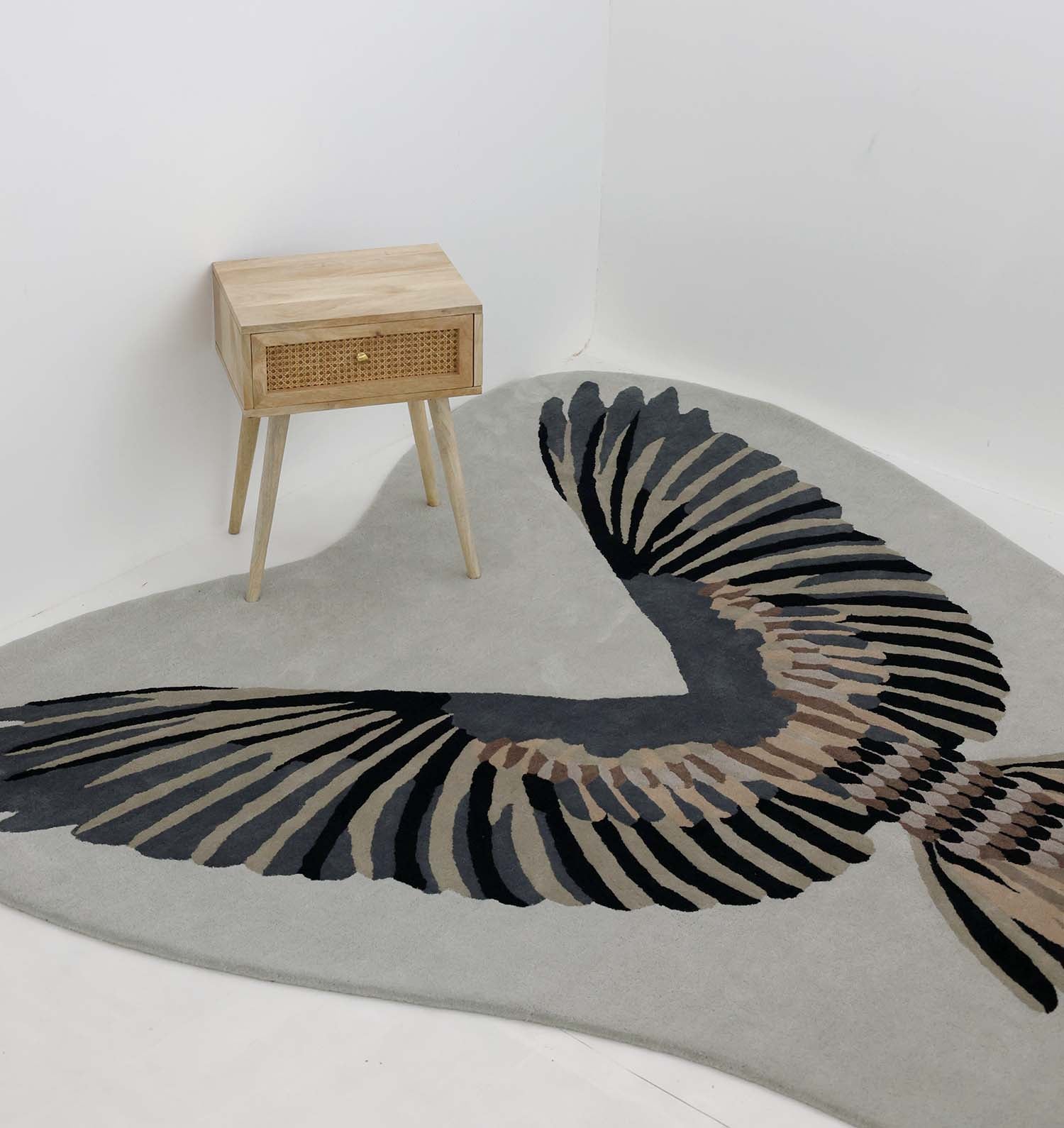 A wooden side table with a wicker drawer stands on the Unshape Handmade Multi Color Rug HTW-82 by RAMSHA HOME, which displays a large bird wing pattern in black, brown, and beige hues, set against a white wall.