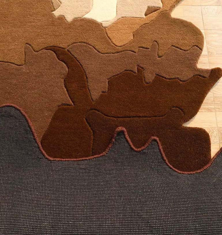 The RAMSHA HOME Handmade Shape Brown Color Area Rug - HTW 86 is a handtufted wool rug with an irregular wavy shape and abstract tan and dark gray patterns, resembling layered earth, displayed on a light wood floor.