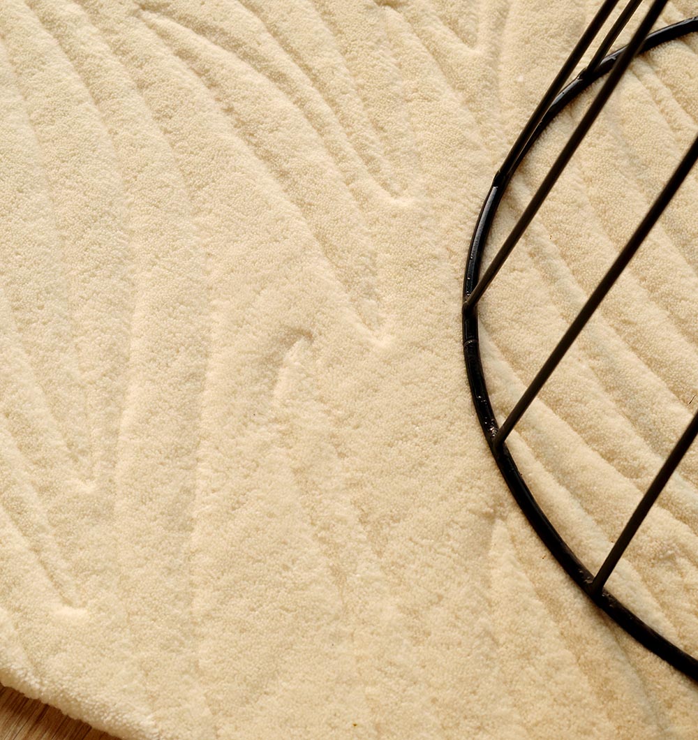 A close-up of the RAMSHA HOME Natural White Color Area Carpet - HTW 88, featuring subtle wave patterns in beige, partially covered by a round black metal wire frame.