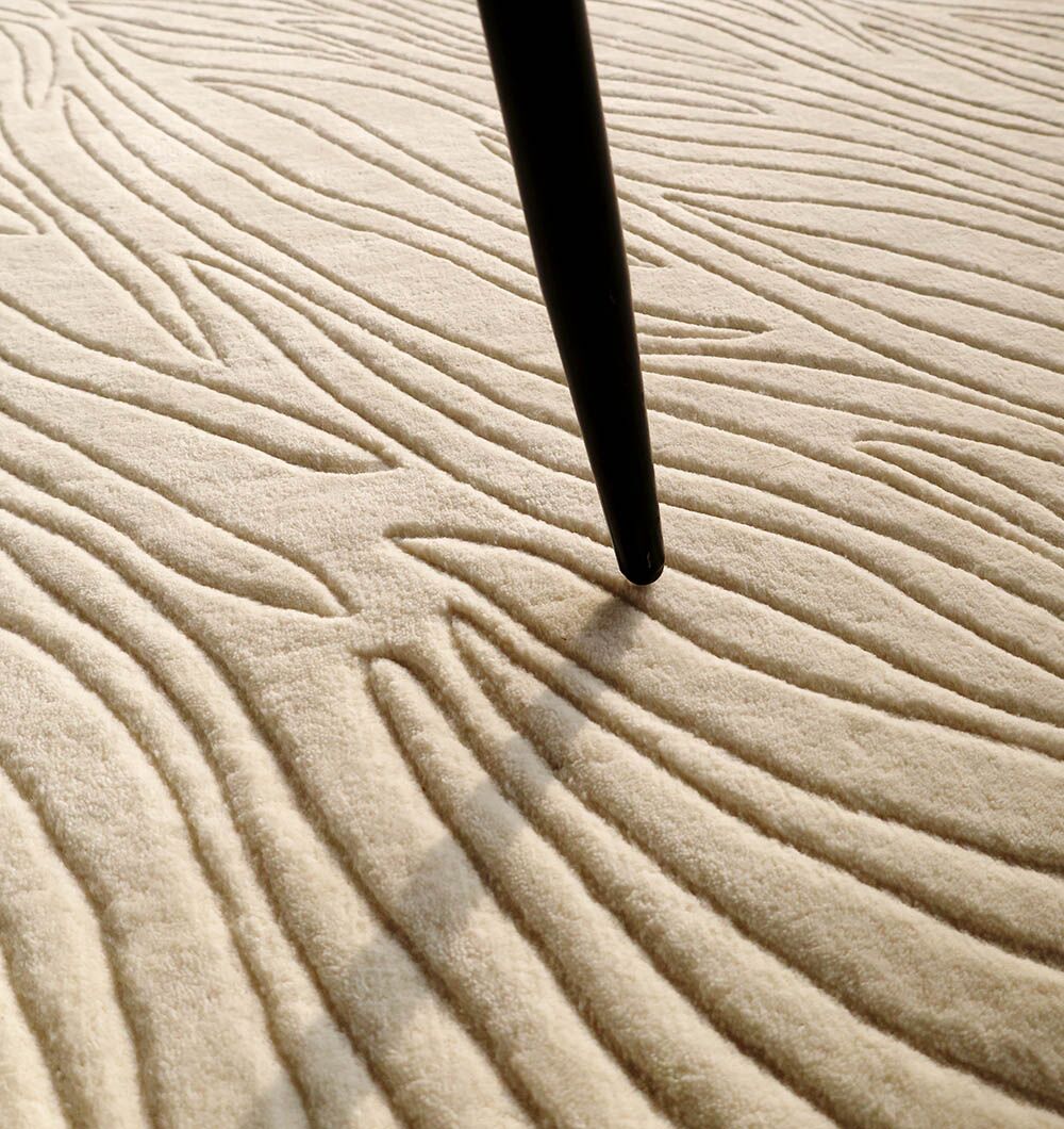 A close-up of the RAMSHA HOME Natural White Color Handmade Area Rug (HTW-89, 300x400 CM) shows its wavy, raised patterns with a black furniture leg resting on its soft, textured surface.