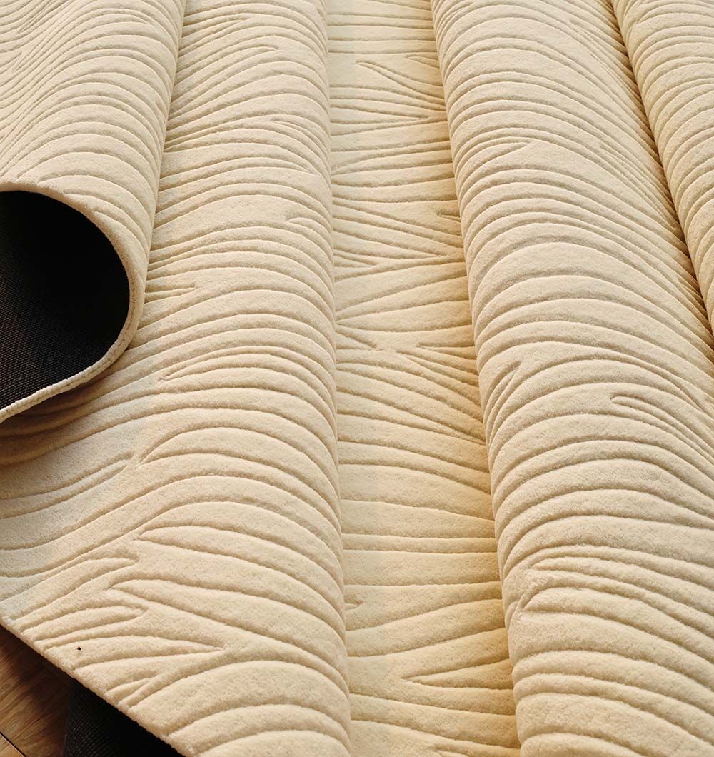 A close-up of the RAMSHA HOME Natural White Color Handmade Area Rug (HTW-89, 300x400 cm) reveals its light beige, raised wavy and linear patterns. The hand-tufted rug is partially rolled to show its thickness and flexible material.