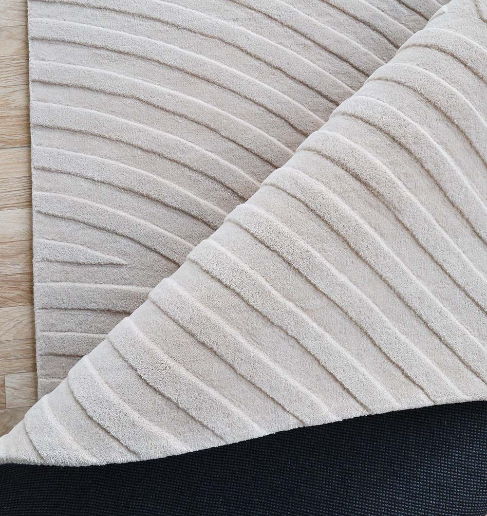 A close-up of the Off White Color Decor Area Rug - HTW-95 (300X400 CM) by RAMSHA HOME shows its hand-tufted, raised wavy pattern, partially lifted to reveal a black non-slip backing on a light wood floor.