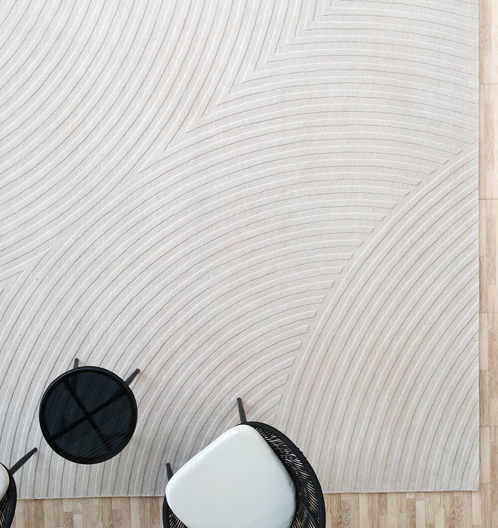 The RAMSHA HOME Off White Color Decor Area Rug - HTW-95 (300x400 CM) features a raised, curved line pattern and is shown on a wooden floor beside two chairs and a small round black table.