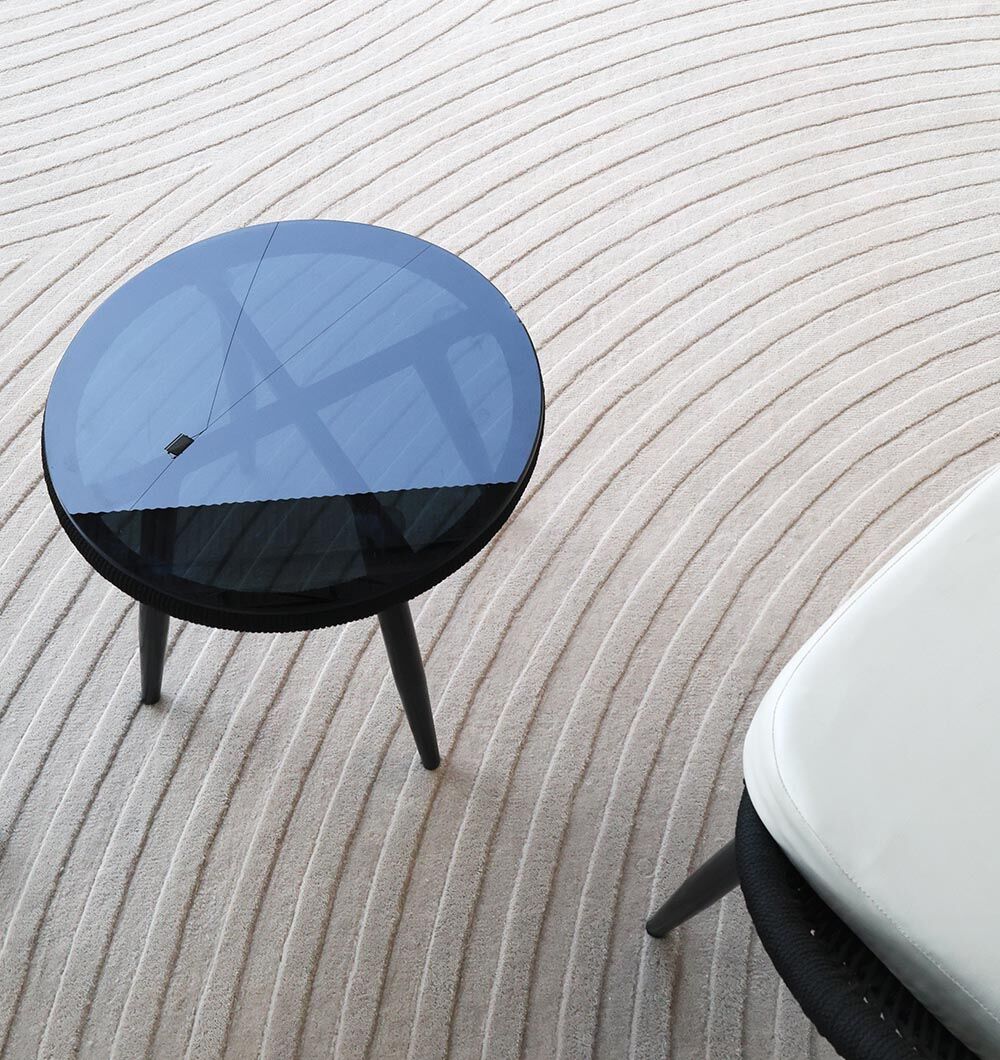 An Off White Color Decor Area Rug - HTW-95 (300X400 CM) by RAMSHA HOME lies beneath a small round black glass-top table and part of a white cushioned chair. The rug features hand-tufted concentric curved lines.