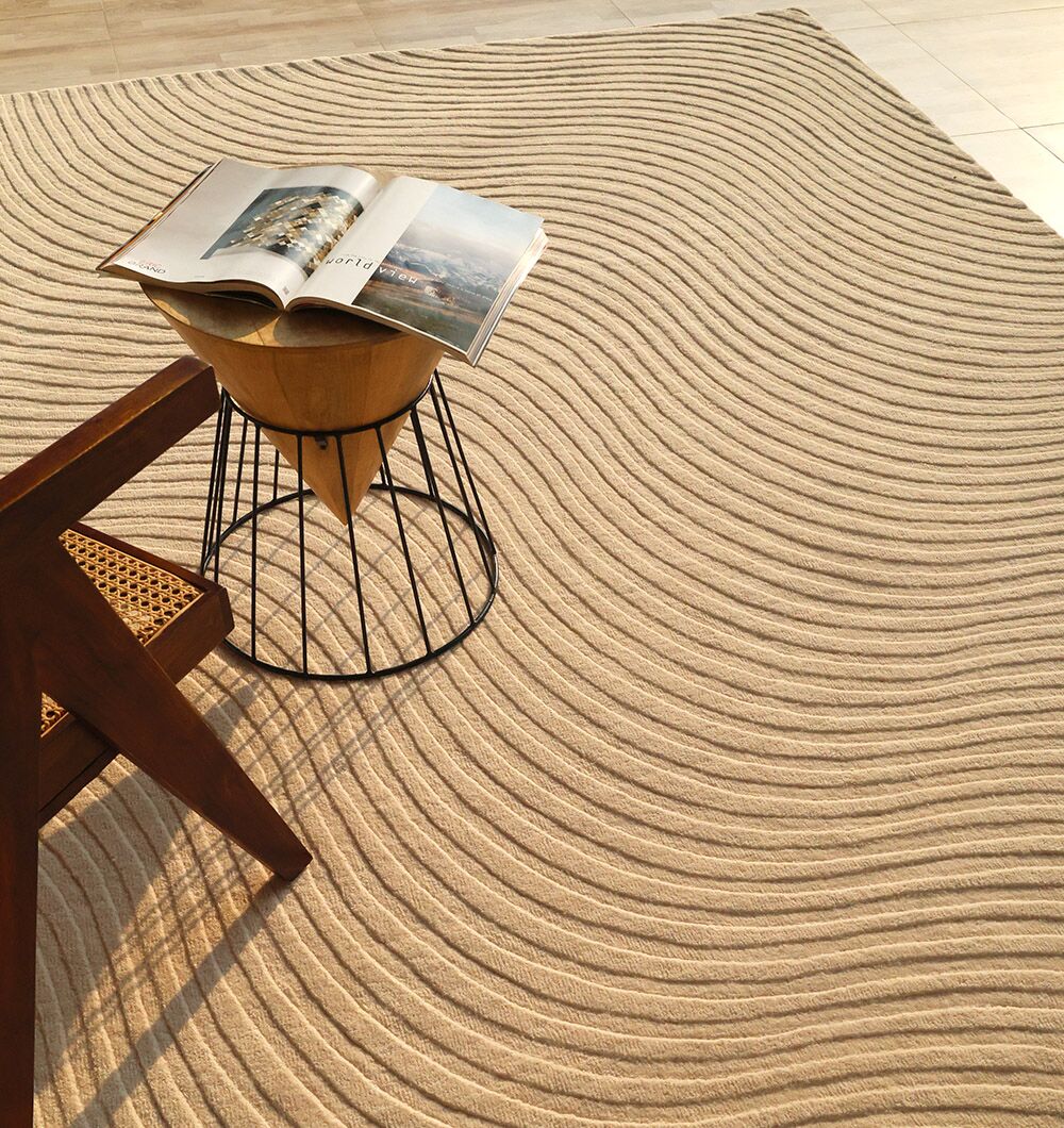 The RAMSHA HOME Home Decor Handmade Area Rug - HTW 96, in beige with wavy textured lines, is placed next to a wooden chair and a round table with an open magazine in a bright, modern room.