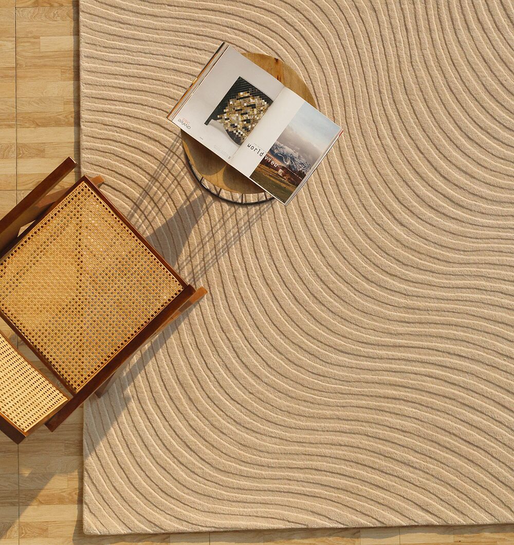 A woven chair and a small round wooden table with an open book sit on the RAMSHA HOME Home Decor Handmade Area Rug - HTW 96, a beige wool rug with raised wavy lines, atop a light wood floor.