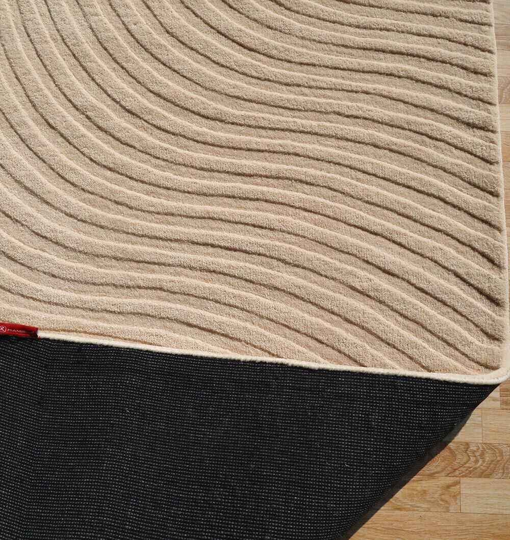 The RAMSHA HOME Home Decor Handmade Area Rug - HTW 96 features beige wool with wavy lines, displayed on a wooden floor. A lifted corner reveals its black non-slip backing.
