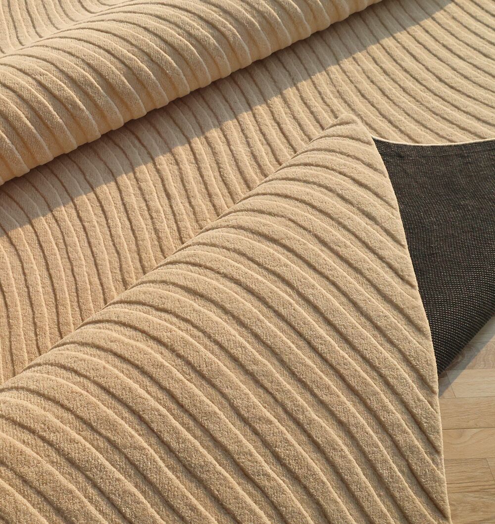 The RAMSHA HOME Home Decor Handmade Area Rug - HTW 96, in beige wool with a wavy pattern, is shown partially rolled to display its textured neutral design and dark non-slip backing on a light wooden floor.