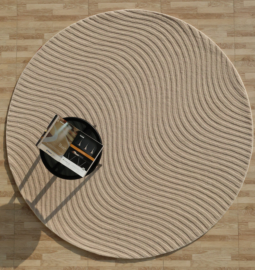 The RAMSHA HOME Home Decor Handmade Area Rug - HTW 96, a round beige wool rug with wavy lines, lies on a light wood floor; a black circular table with an open magazine rests partly on the textured neutral rug, casting a soft shadow.