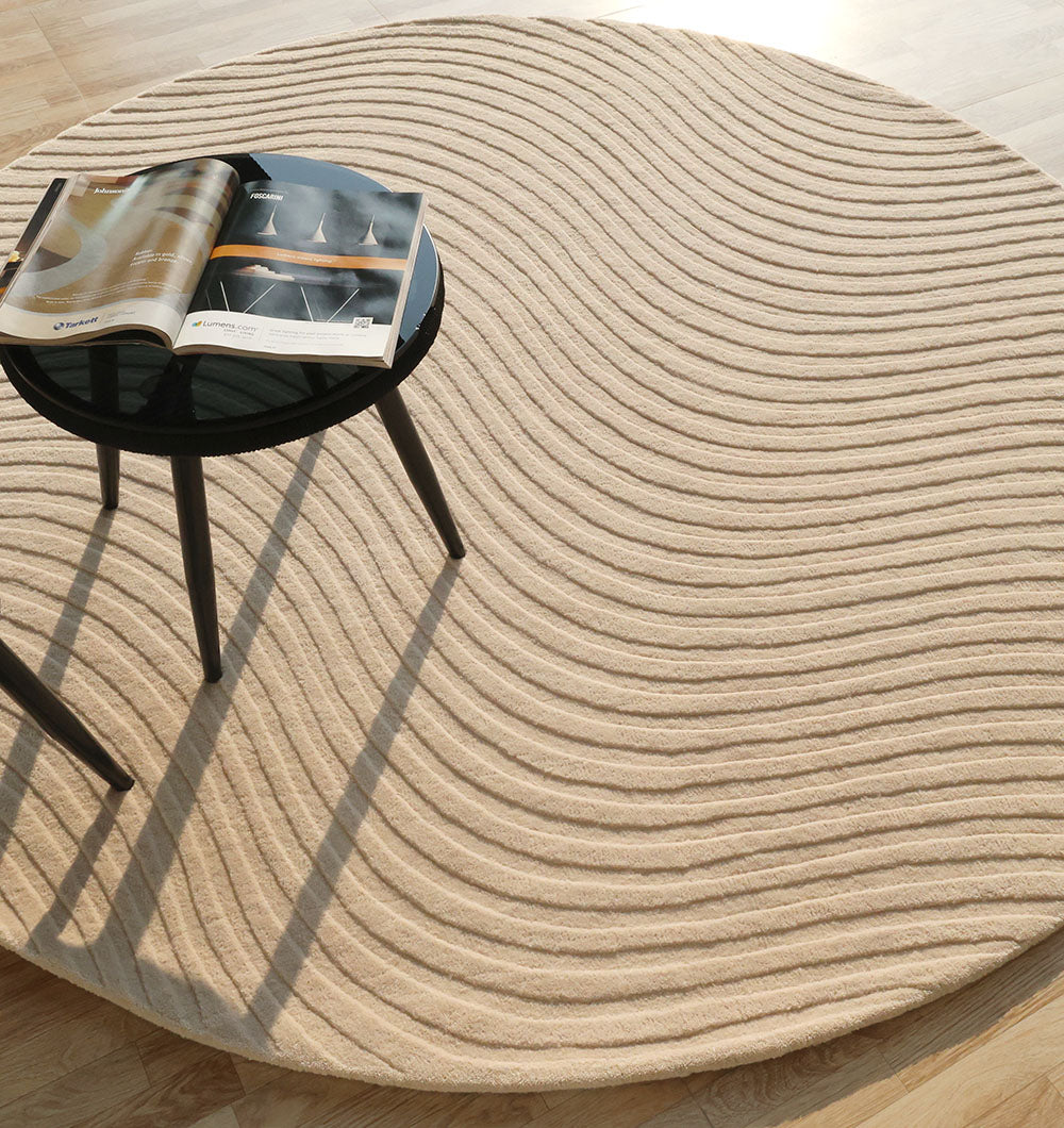 The RAMSHA HOME Home Decor Handmade Area Rug - HTW 96, a round beige wool piece with wavy textured lines, sits on a light wooden floor beneath a small black three-legged table holding an open magazine in sunlit surroundings.
