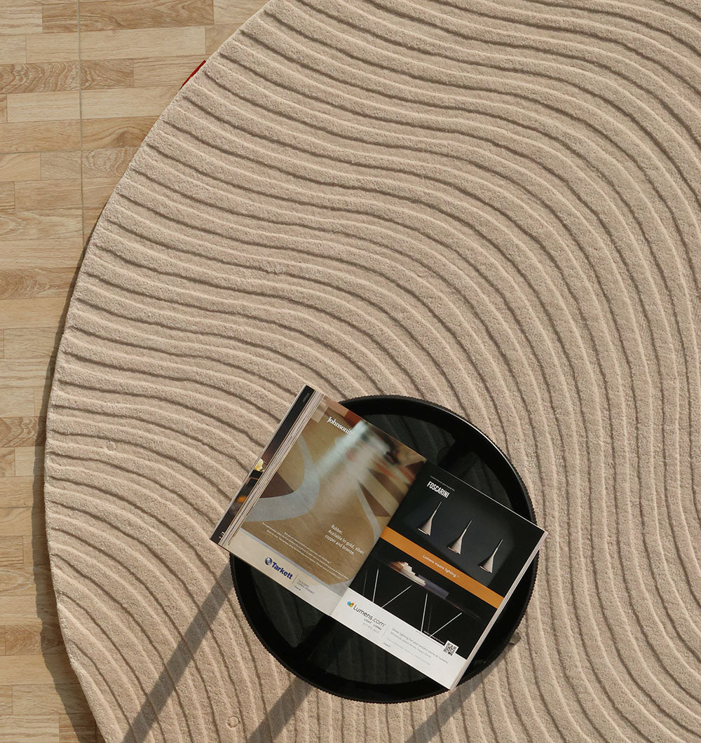 The RAMSHA HOME Home Decor Handmade Area Rug - HTW 96, featuring a circular textured neutral design with wavy lines, lies on a light wood floor. Atop the rug, a round black tray holds an open magazine showing modern interiors.