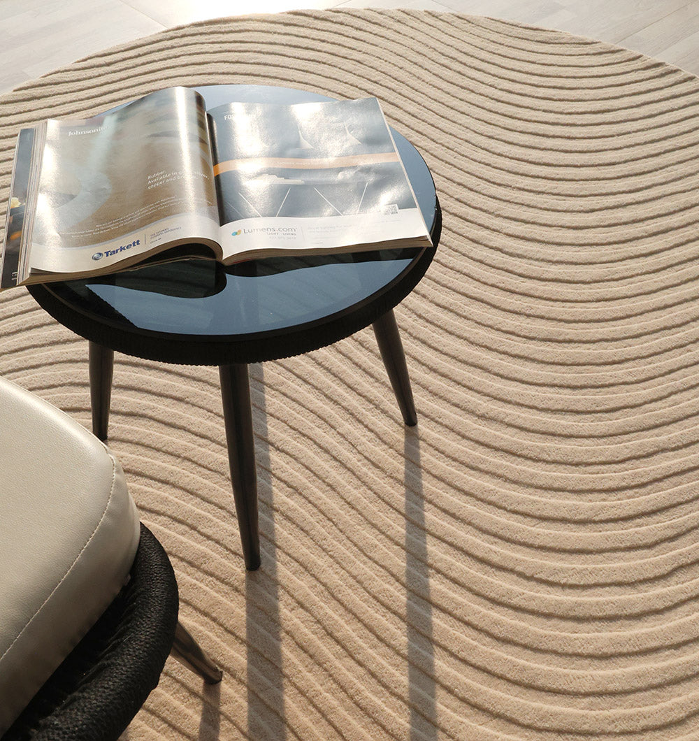 A round glass-top table with three dark legs holds an open magazine. The table rests on the RAMSHA HOME Home Decor Handmade Area Rug - HTW 96, which features wavy textures. Part of a light chair and sunlight casting soft shadows are also visible.