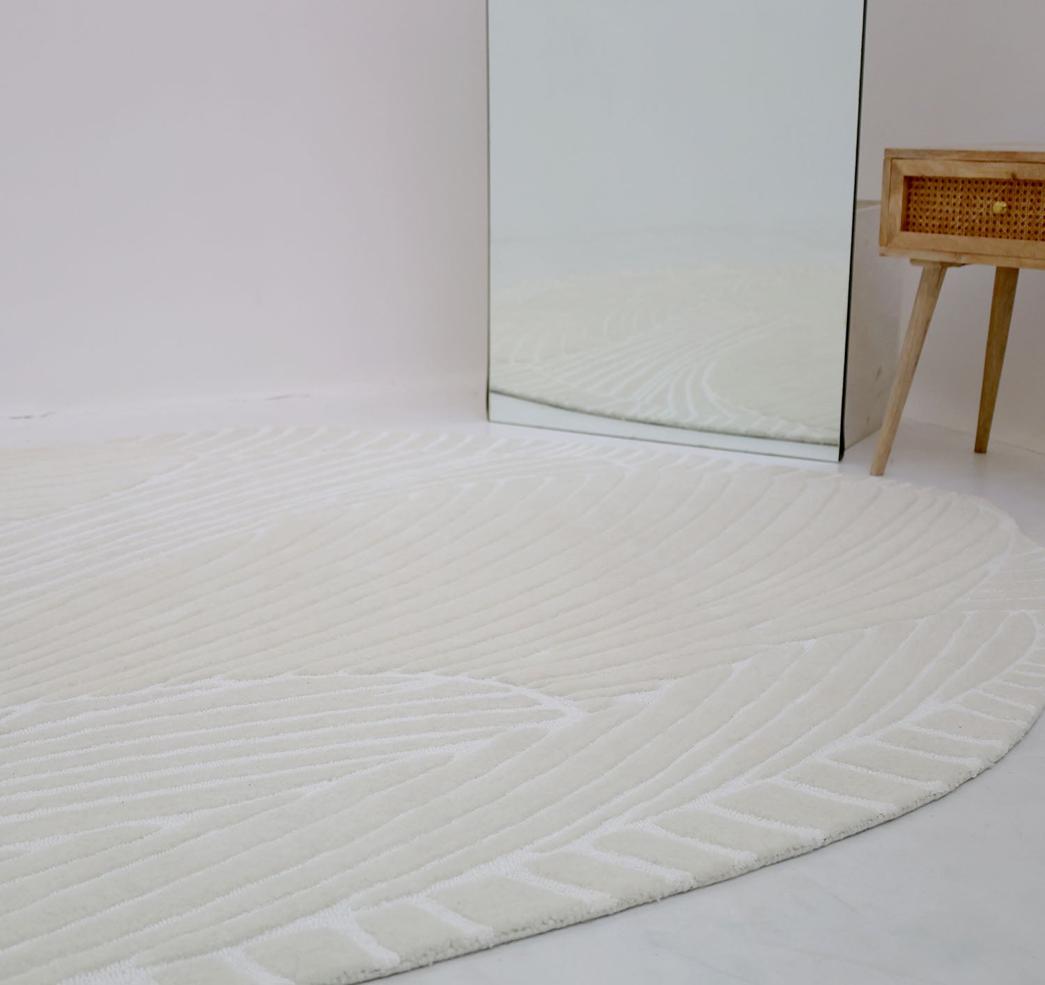 The RAMSHA HOME Handmade Carpet 200x300 cm Shaped Area Rug HTWV-103, cream-colored with subtle patterns, is placed near a large mirror and a small wooden table with a woven front in a minimal, modern setting.