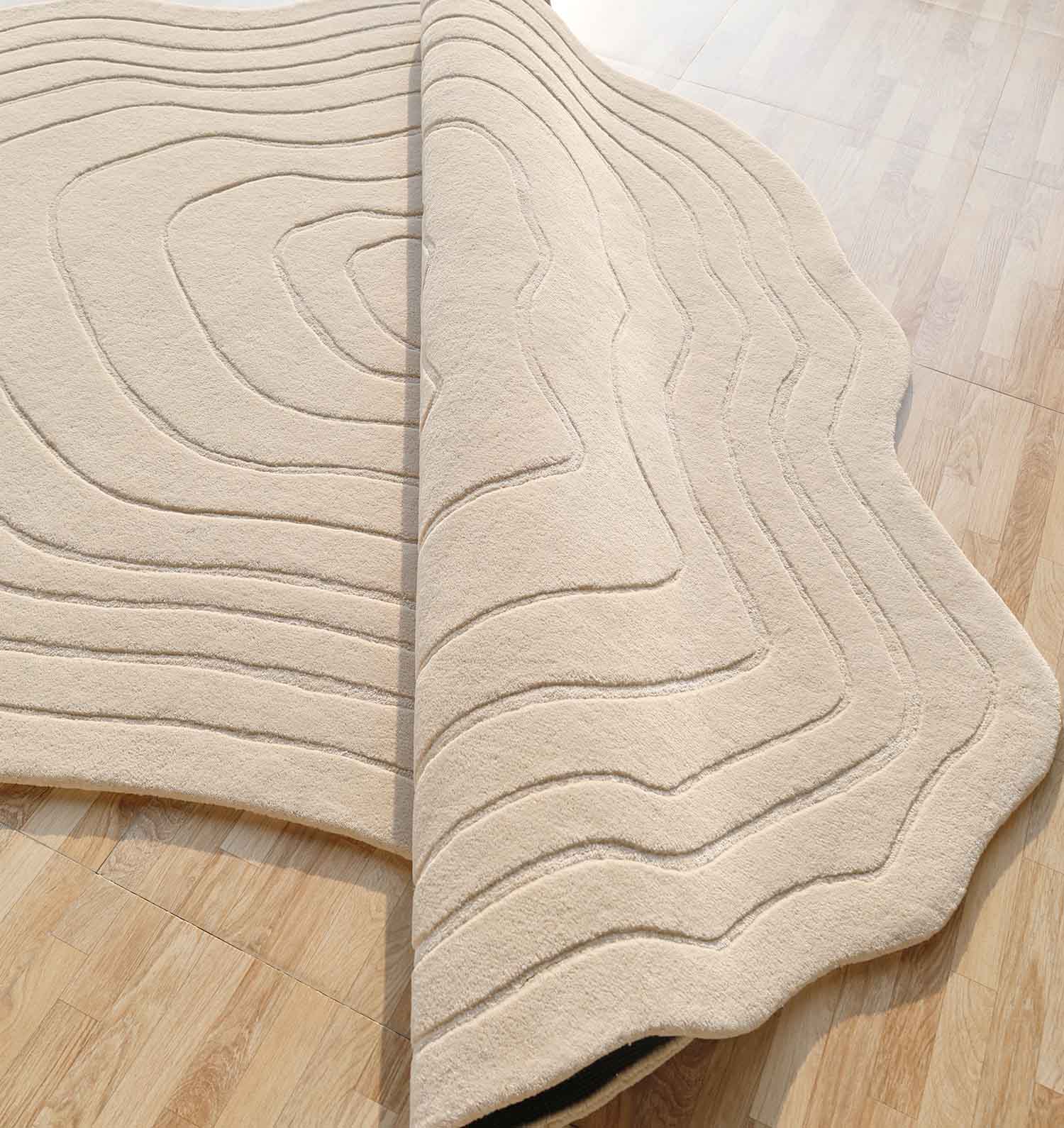 The Hand Tufted Beige Color Decor Rug HTWV-108 by RAMSHA HOME features raised, wavy lines and a modern texture in a cream hue. It is shown partially folded back to reveal wood-patterned flooring beneath, with soft lighting highlighting its details.