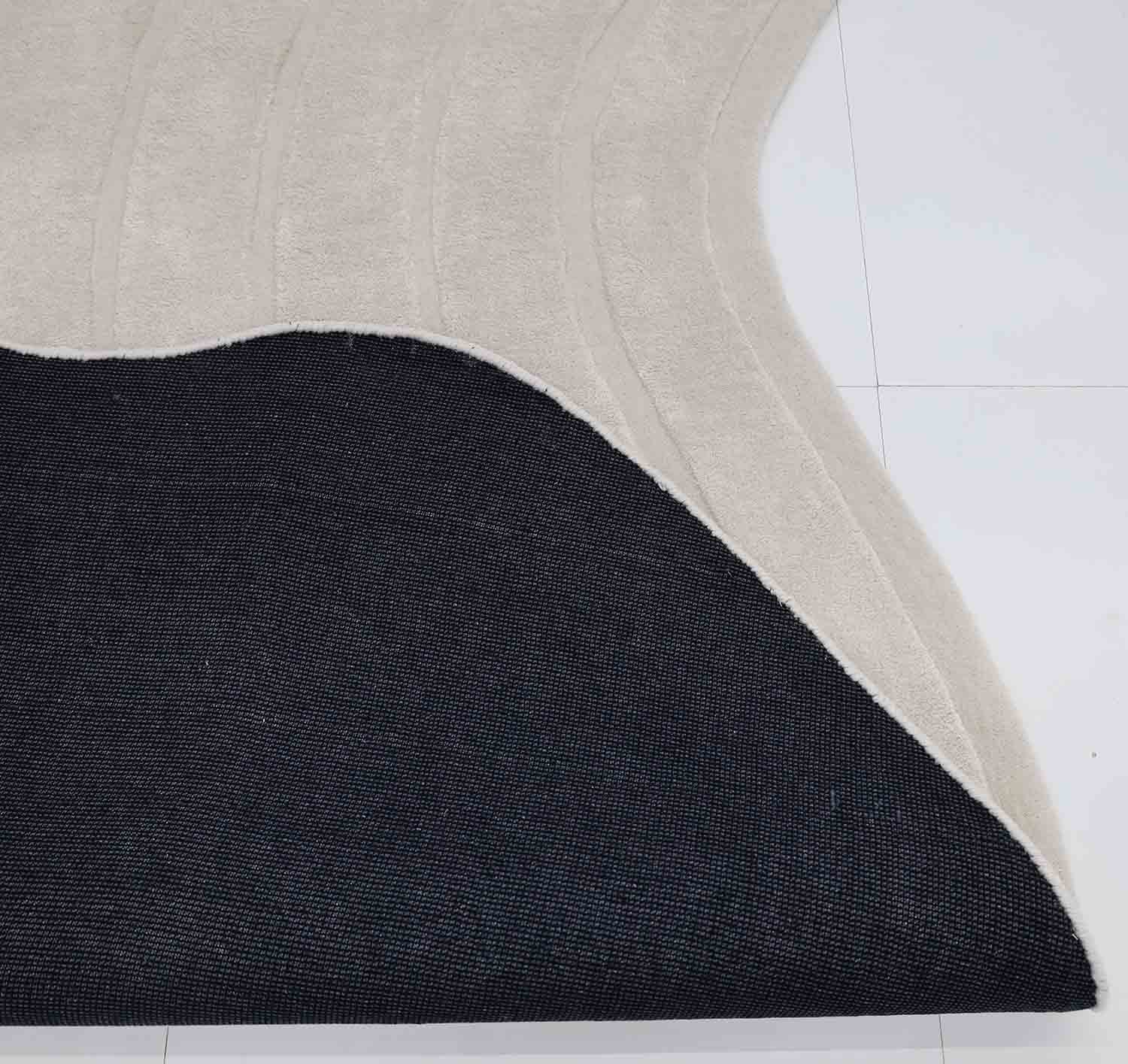A close-up of the RAMSHA HOME Luxurious Off-White Hand-Tufted Rug (HTWV 111, 200x300cm) shows its black non-slip backing, partially lifted to reveal the underside on a white tiled floor.