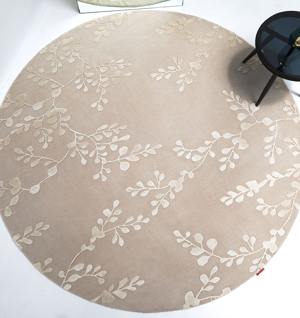 The RAMSHA HOME Artisan Handmade Decorative Area Rug – HTWV-116 is a round wool rug in beige with a cream botanical pattern. A small black glass-top side table sits at its edge, and part of a mirror appears in the background.