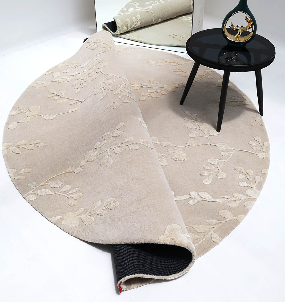The RAMSHA HOME Artisan Handmade Decorative Area Rug – HTWV-116, a light beige hand-tufted round wool rug with a subtle leaf pattern, is partially folded under a black round side table beside a mirror reflecting the scene.