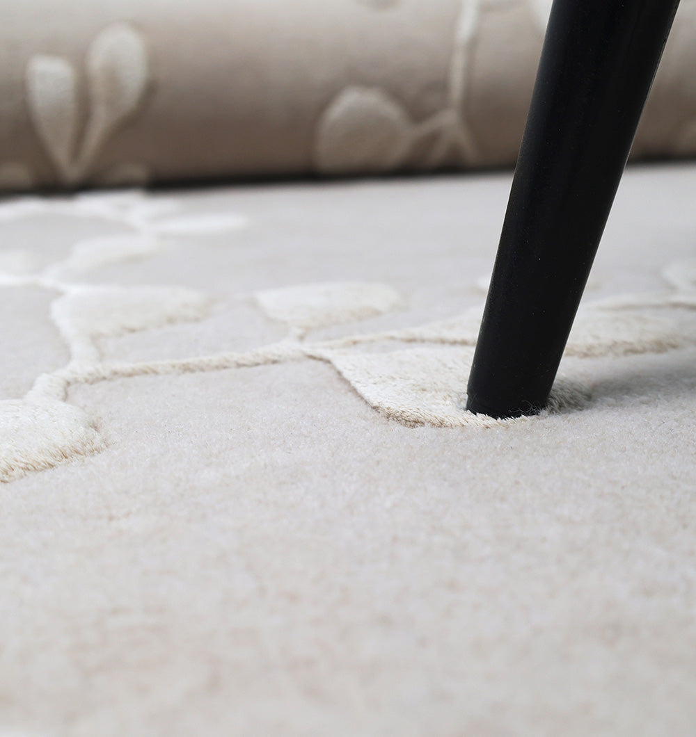 Close-up of a black furniture leg pressing into the RAMSHA HOME Artisan Handmade Decorative Area Rug – HTWV-116 in light beige, showing the subtle floral pattern and textured indentation.