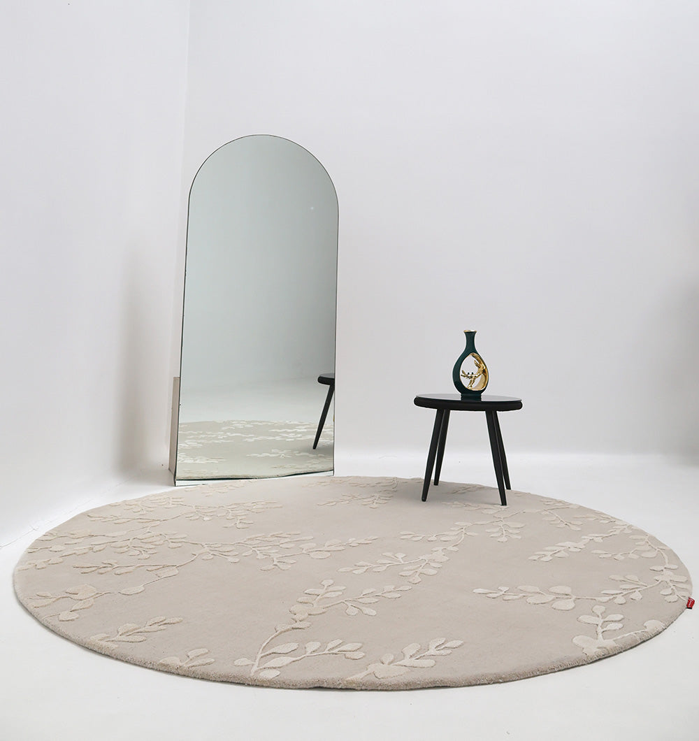 The RAMSHA HOME Artisan Handmade Decorative Area Rug – HTWV-116 with a subtle leaf pattern is placed in a minimal room. A tall arched mirror and a small black table with a vase rest on the rug, set against a plain white background.