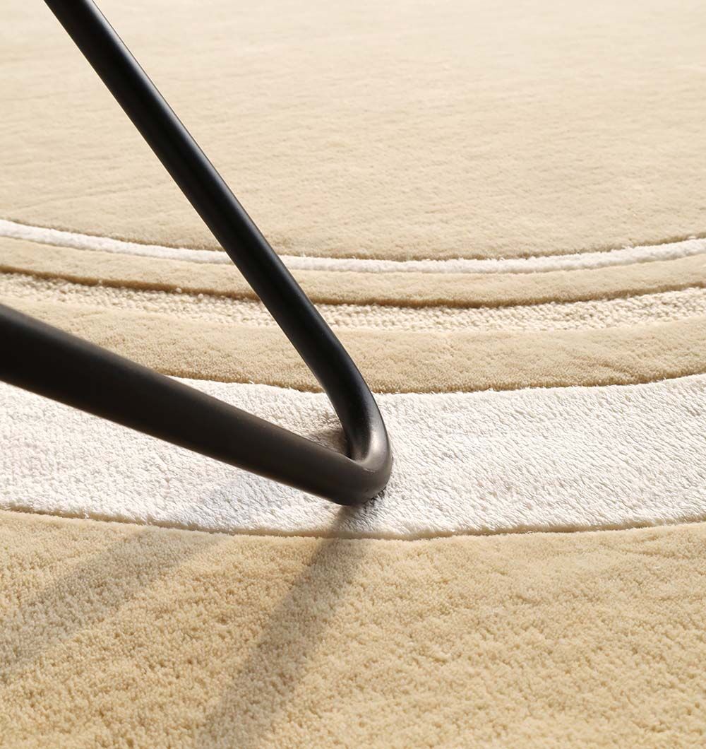 A close-up of a black metal table leg on the RAMSHA HOME Multi Color Handmade Rug - HTWV-120 shows beige and white handtufted wool with subtle raised patterns. Sunlight softly highlights this modern rug’s textured, circular design.
