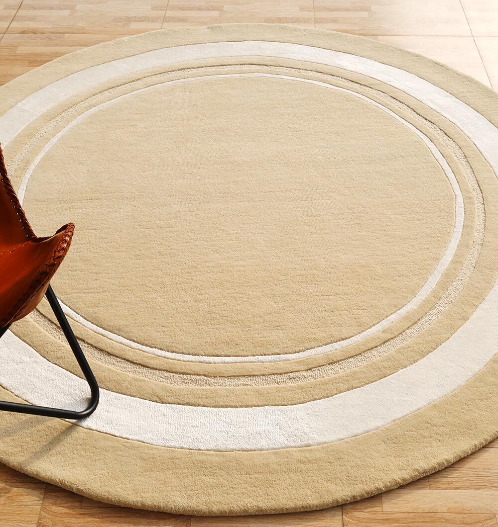 The RAMSHA HOME Multi Color Handmade Rug - HTWV-120, with concentric beige patterns in light and dark tones, is handtufted in wool and placed on a wooden floor, with a brown leather chair edge visible for added warmth.