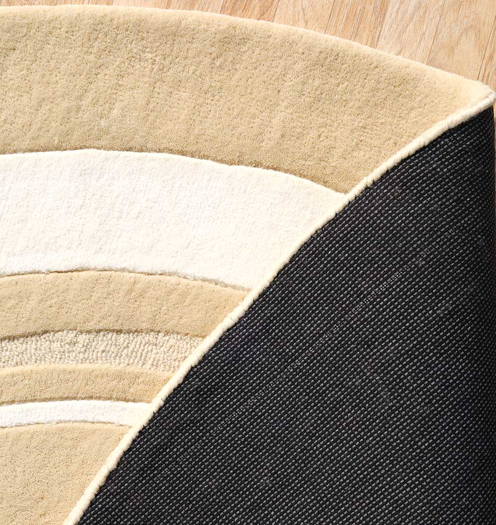 A close-up of the RAMSHA HOME Multi Color Handmade Rug - HTWV-120 in beige and cream with curved stripes, partially lifted to show its dark textured non-slip backing on a light wood floor.