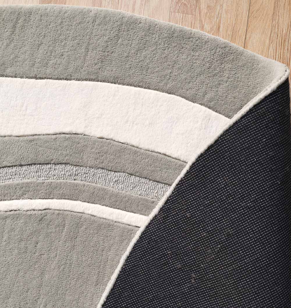 A close-up of the RAMSHA HOME Grey & White Color Round Handmade Rug - HTWV-121 on a wooden floor, featuring gray, white, and dark gray curved stripes with its textured underside partially flipped over.