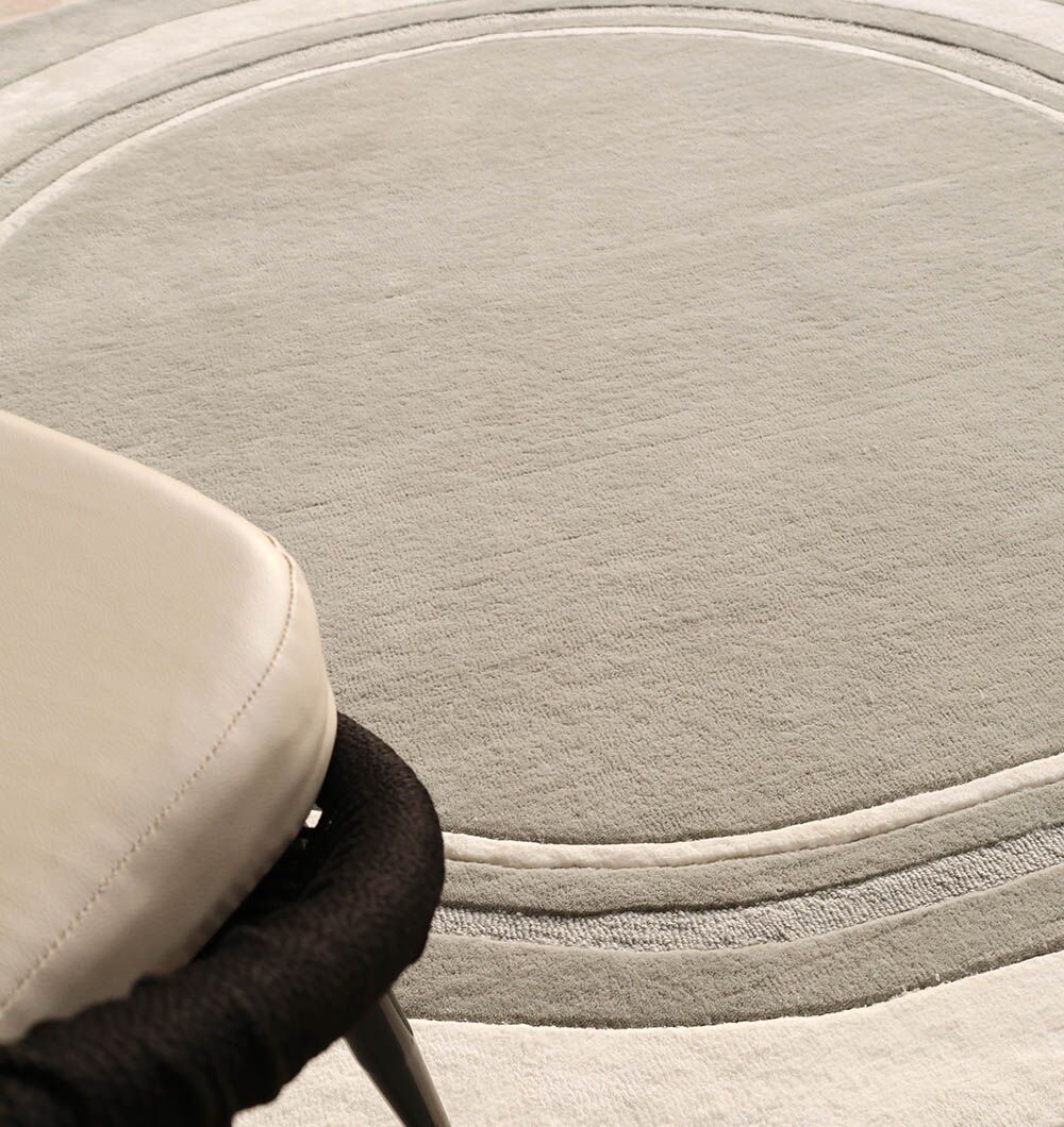 A close-up of the RAMSHA HOME Grey & White Color Round Handmade Rug - HTWV-121, featuring handtufted concentric rings, partially reveals a light cushioned chair with black legs in the lower left corner.