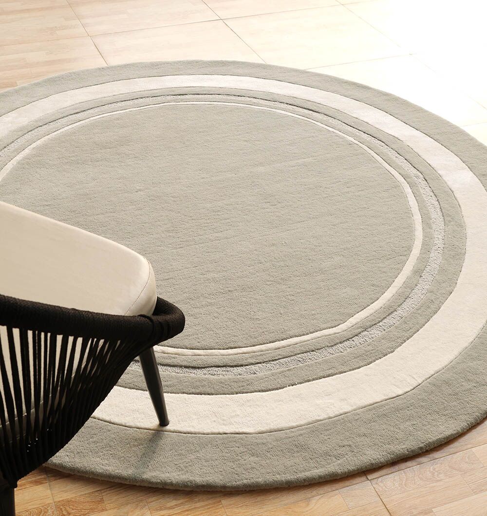 The RAMSHA HOME Grey & White Color Round Handmade Rug - HTWV-121, featuring light gray with concentric white and dark gray rings, is displayed on a light wood floor. A modern black-framed chair with a white cushion is partially visible on the left.