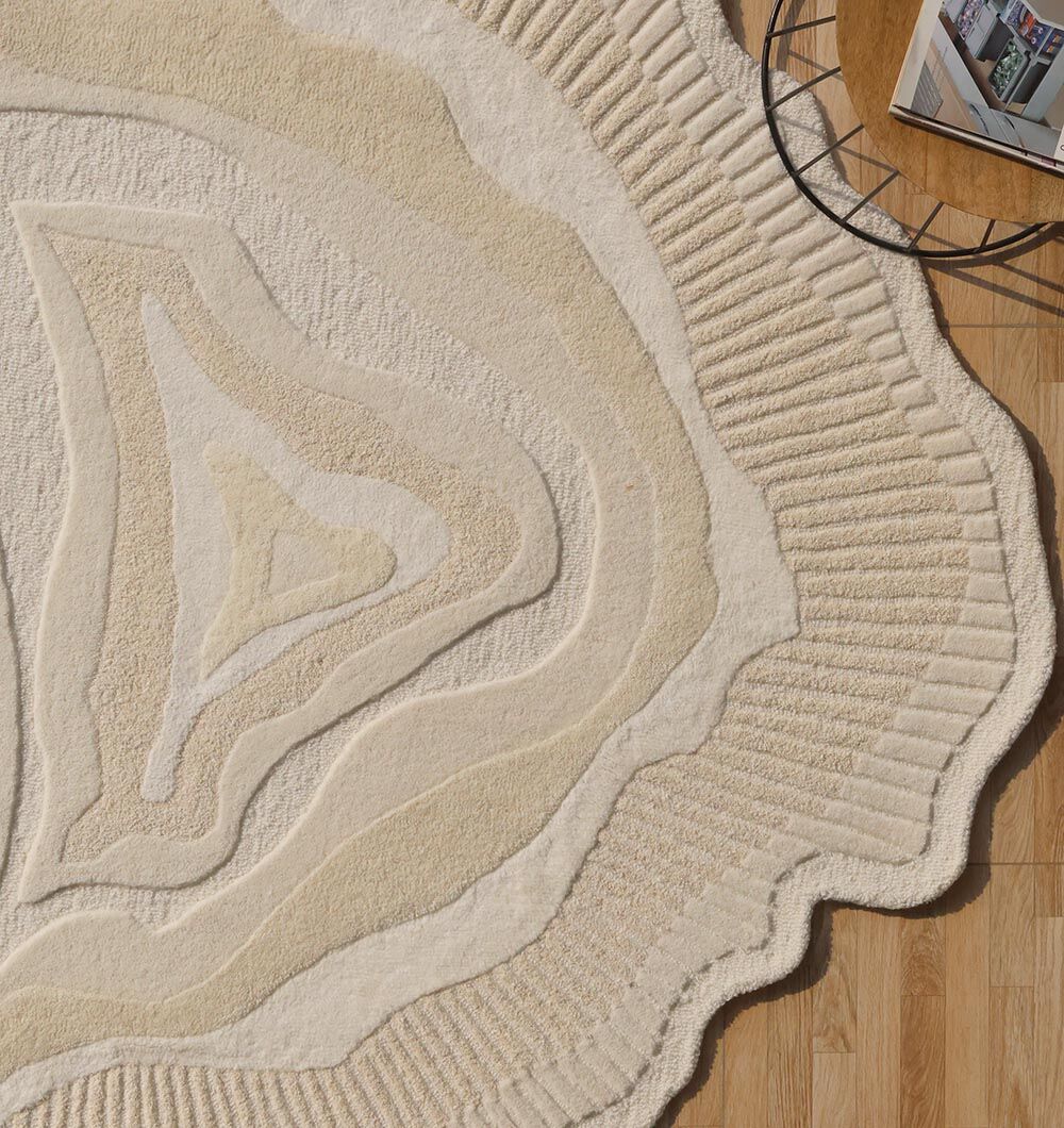 The RAMSHA HOME Natural Handmade Area Rug - HTWV-126 in cream and beige with wavy layers accents a wooden floor. In the upper right, a round black metal table with stacked magazines is partially visible.