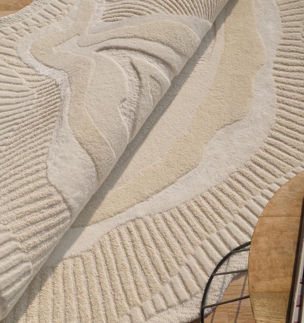 A close-up of the RAMSHA HOME Natural Handmade Area Rug - HTWV-126 in beige and cream, featuring raised wavy patterns, partially rolled on a wooden floor near a round table and black metal chair.